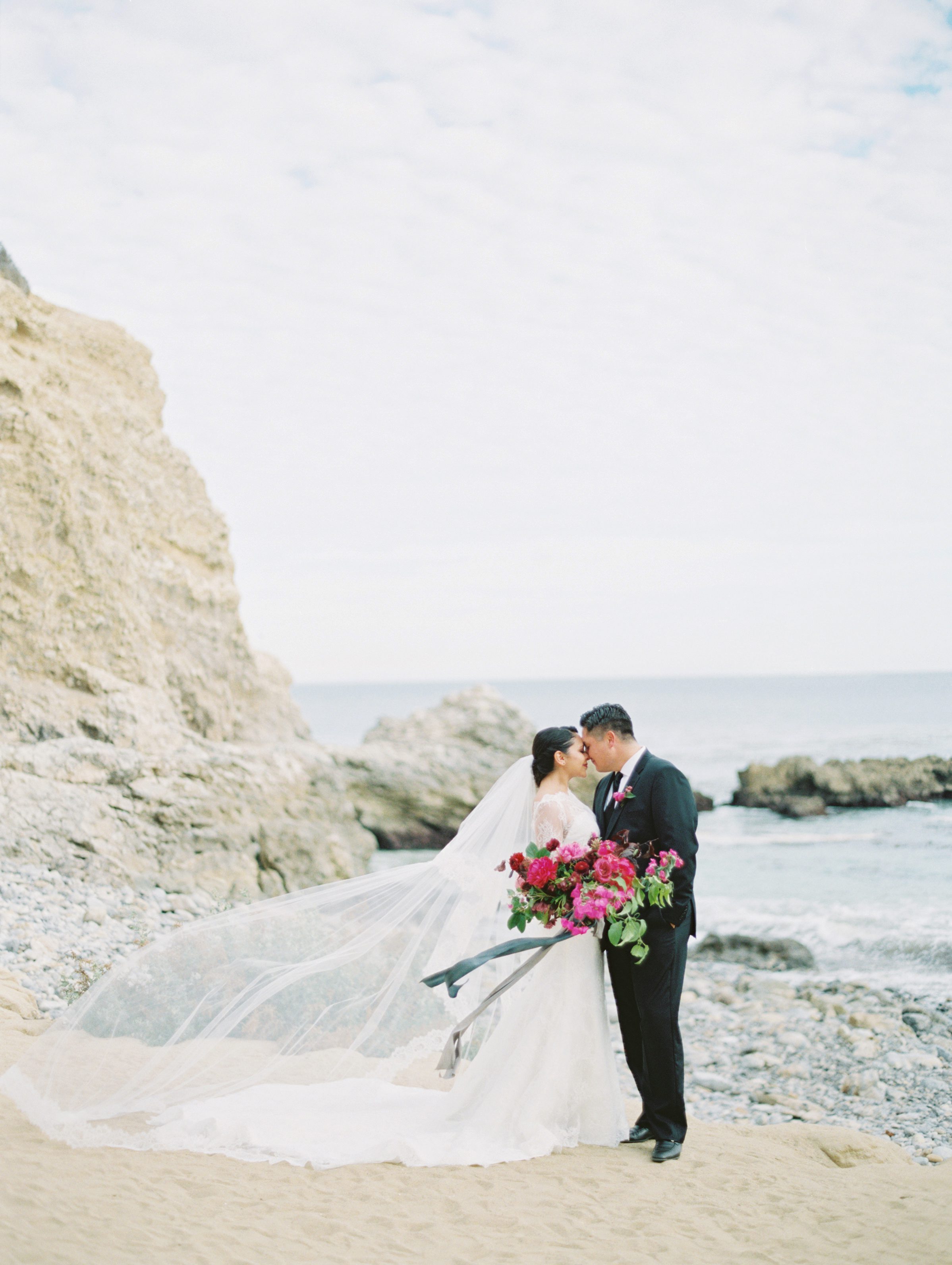 Bride and groom on ocean rock with bouquet and blowing veil